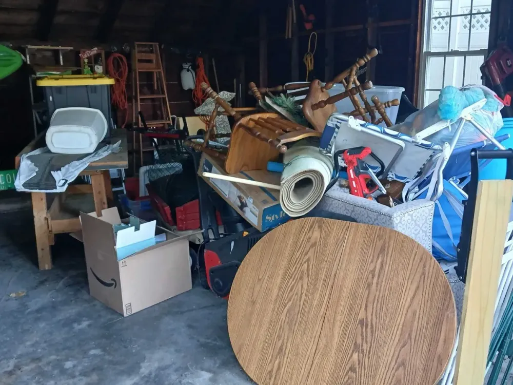 A packed suburban garage full of chairs, a table top, rolled carpet, an armchair and folding lawn chairs before a cleanout