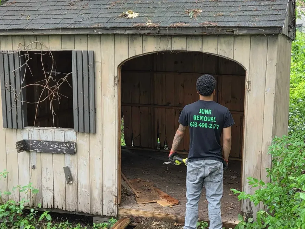 A Trash Can Willy's crew member with a reciprocating saw standing at the open door of an emptied backyard shed