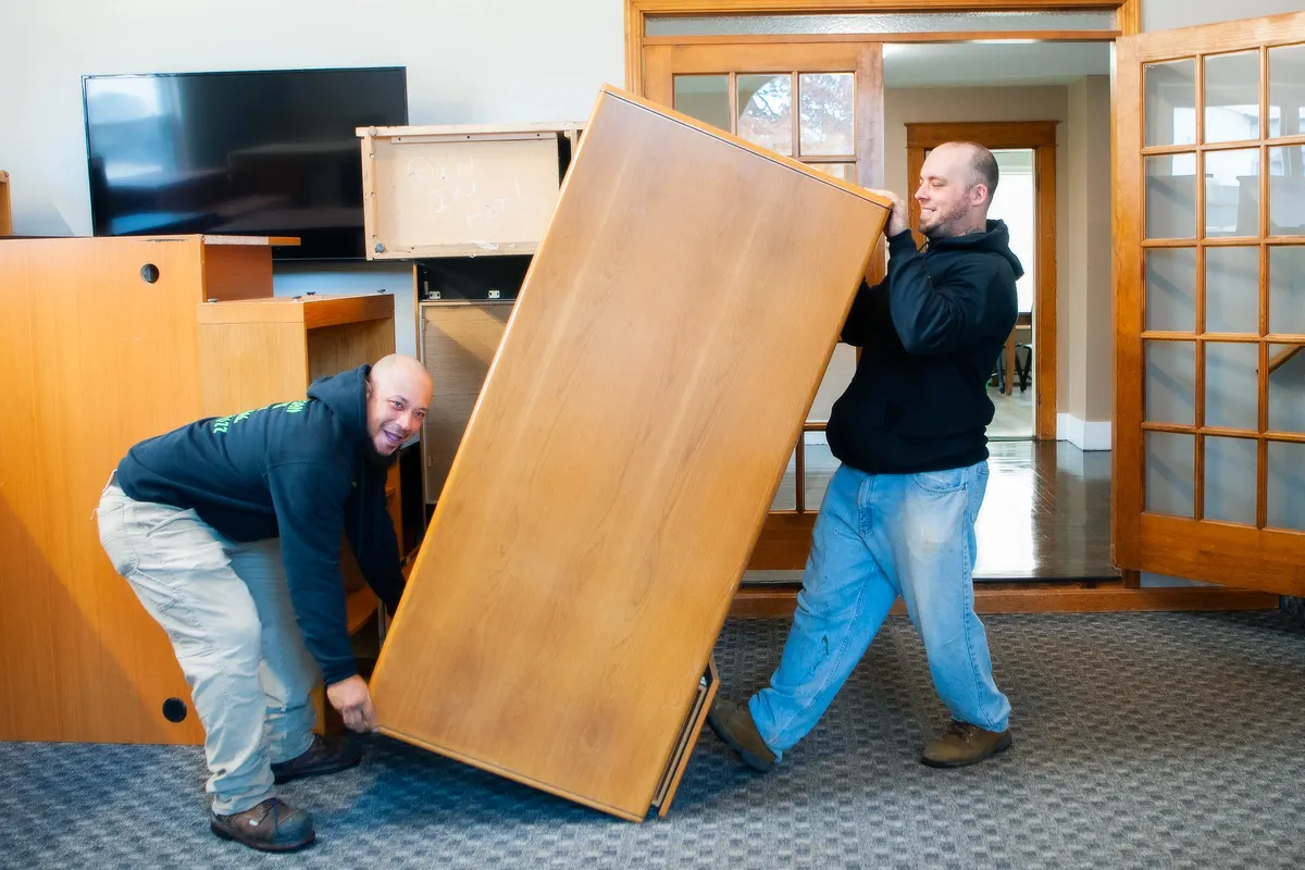 Two Trash Can Willy's crew members carrying a tall dresser out through a doorway