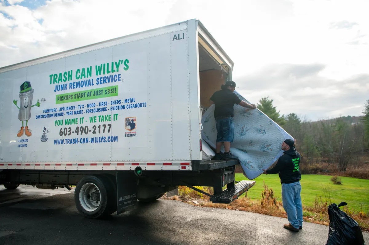 Two crew members lifting a mattress onto the liftgate at the back of a Trash Can Willy's box truck