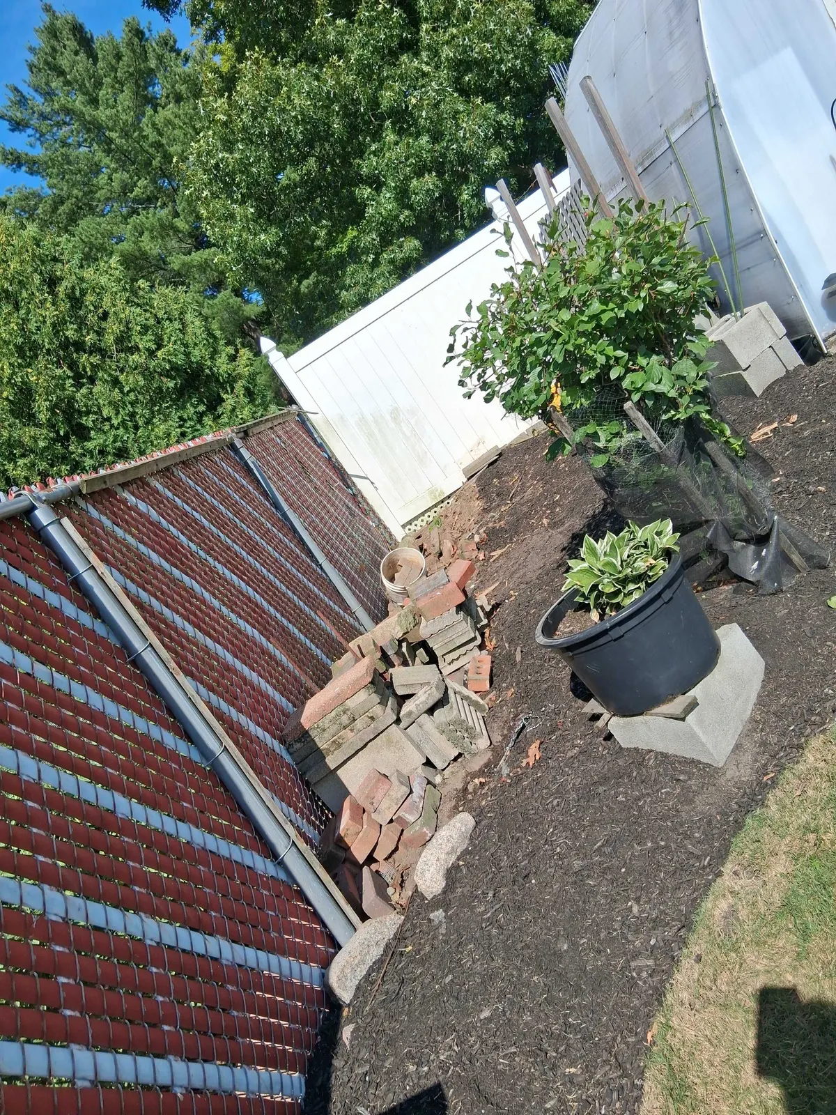 An old chain link fence with privacy slats leaning over, with bricks and pavers stacked along the line