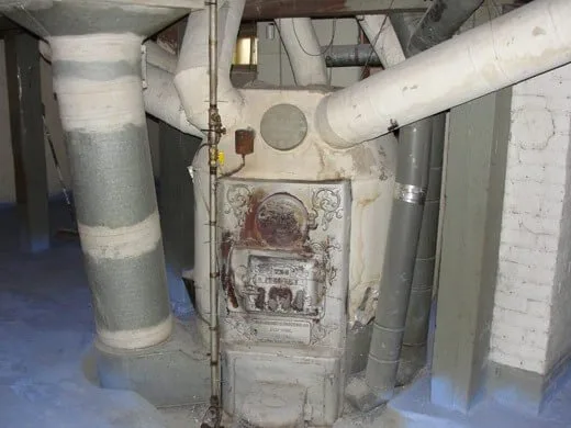 An old cast-iron furnace standing in a basement, with insulated ducts running off it and a concrete floor around it