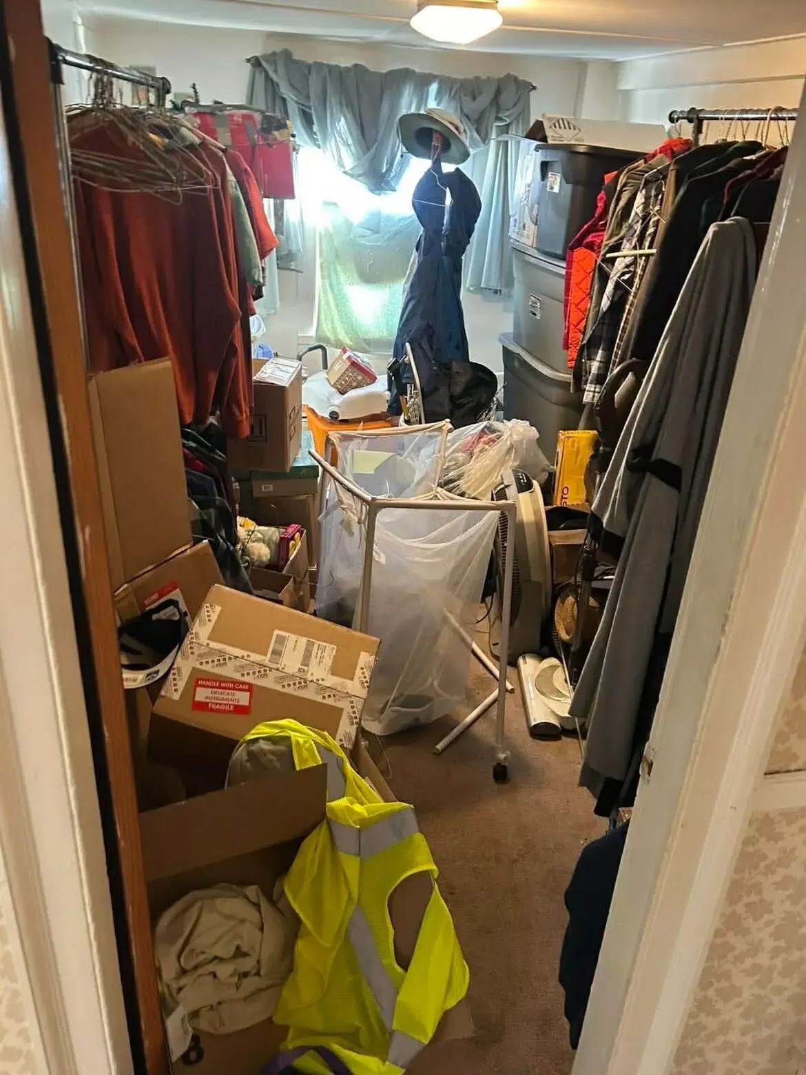 A packed bedroom with clothes racks and boxes to the door, before a cleanout